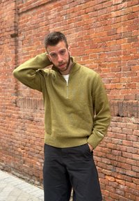 Homme aux cheveux courts et barbe, portant un pull à col zippé vert olive et un pantalon noir, debout avec une main sur la nuque près d'un mur en briques.