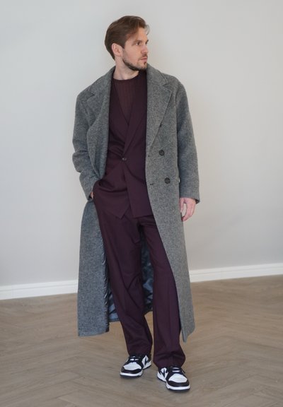 Abrigo de lana gris con un diseño de doble botonadura, combinado con un traje burdeos profundo y zapatillas en blanco y negro, sobre un suelo de madera.