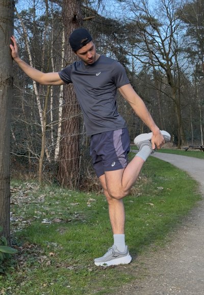 Conjunto atlético que incluye una camiseta gris oscura y pantalones cortos azul marino. Corredor estirándose junto a un árbol, con zapatillas gris claro y calcetines cortos.