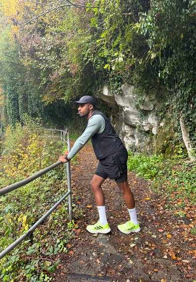 Hombre con ropa deportiva apoyado en una barandilla en un sendero frondoso del bosque junto a una pared de roca durante el otoño.