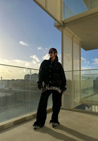 Sort denimjakke med store knapper og kontrasterende syninger, parret med sorte bukser med hvid "Mansion" tekst og spidse sorte højhælede sko.