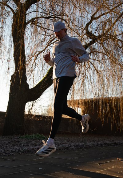 Hombre trotando al aire libre por un camino pavimentado al amanecer, con gorra, chaqueta gris, leggings negros y zapatillas blancas de correr.