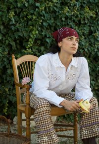 Femme portant un bandana rouge et un pantalon à motifs, assise sur une chaise en bois, en train d'éplucher une pomme, avec un fond de lierre vert.