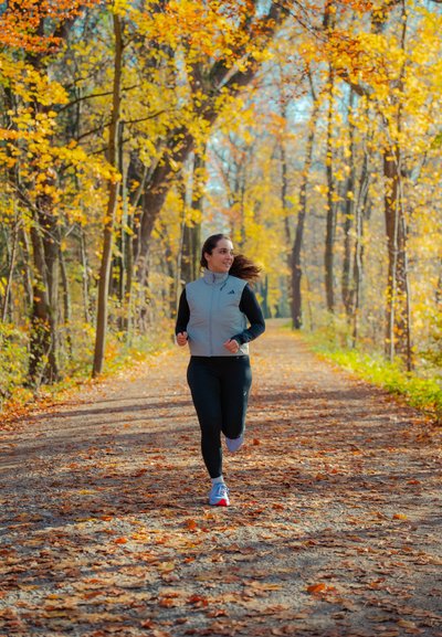 Conjunto deportivo que incluye un chaleco gris claro con cremallera y leggings negros; corriendo por un camino cubierto de hojas, rodeado de árboles amarillos y naranjas.