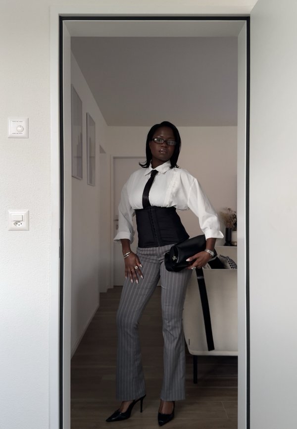 Kvinde iført briller, hvid skjorte, sort slips, korset, stribede bukser og hæle, holder en sort clutch i døren til et moderne rum.