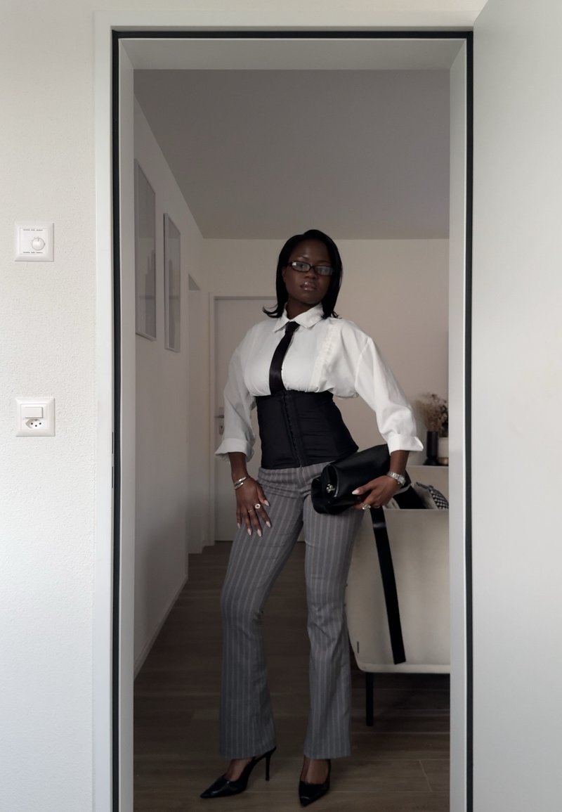 Kobieta w okularach, białej koszuli, czarnym krawacie, gorsecie, paskowanych spodniach i szpilkach, trzymająca czarną kopertówkę w drzwiach nowoczesnego pokoju.