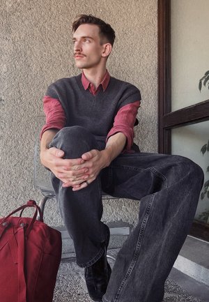 Jean de pierna ancha gris oscuro, camisa roja con cuello bajo un chaleco azul marino. Sentado en una silla transparente, con una bolsa roja descansando cerca. Fondo texturizado.