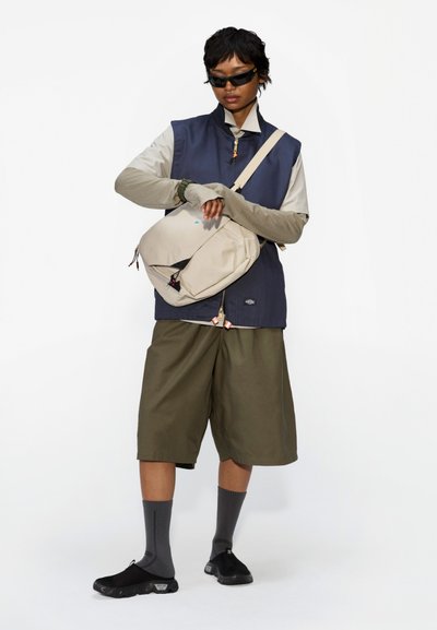Bolso cruzado beige con cremallera, chaleco azul marino sobre camisa de manga larga clara, pantalones cortos oliva, calcetines grises y zapatos negros. Detalles de accesorios presentes.