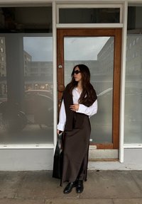 Rochie maxi din satin maro, cămașă albă cu nasturi, ochelari de soare negri și bocanci negri masivi. Ținând o geantă mare neagră. Fundal cu ușă din lemn.