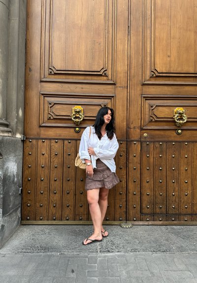 Puertas dobles de madera con anillas en forma de cabeza de león y tachuelas de metal. El sujeto lleva una camisa blanca de mangas largas, una falda marrón en capas y sandalias.