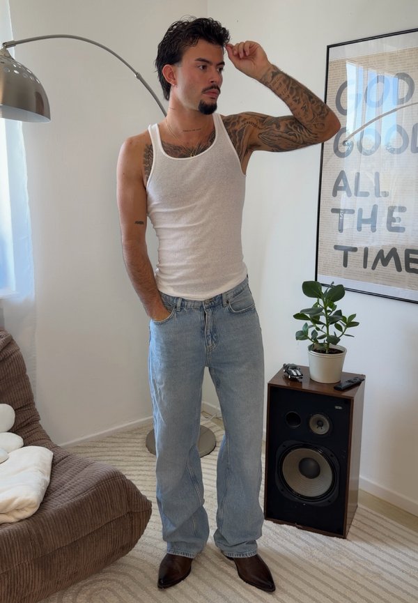 Mand med tatoveringer iført hvid tanktop og jeans står med den ene hånd i lommen i et lyst rum ved siden af en højttaler og en plante.