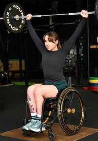 Jeune femme en fauteuil roulant soulevant une barre au-dessus de sa tête dans une salle de sport, portant un haut noir et un short turquoise, avec un tatouage floral sur la jambe gauche.