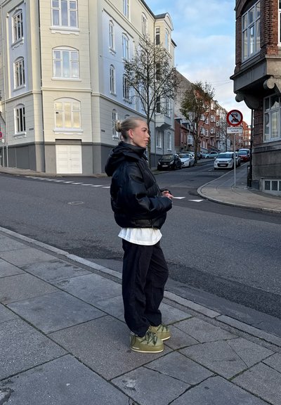 Persona con chaqueta y pantalones negros, zapatos verdes, de pie en una acera en una esquina de la calle con coches aparcados y edificios al fondo.
