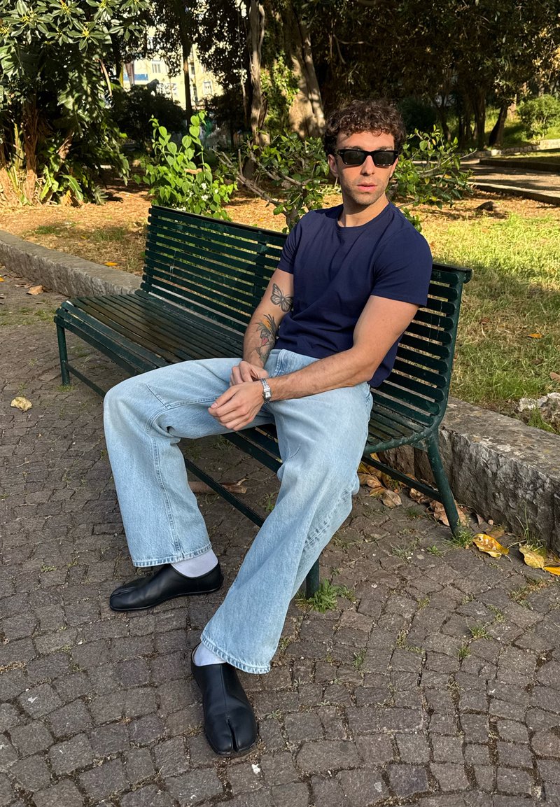 Homme avec des lunettes de soleil et des tatouages assis sur un banc vert dans un parc, portant une chemise bleu marine, un jean clair, des chaussettes blanches et des chaussures noires à orteils fendus.