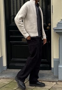 Cardigan gris clair sans col avec poignets côtelés, porté sur une chemise foncée et un pantalon noir à jambes larges, assorti à des chaussures noires épaisses.