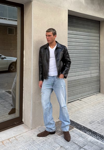 Joven con chaqueta de cuero negra, camiseta blanca, vaqueros claros rasgados y botas marrones, de pie en una acera de la ciudad cerca de una puerta de garaje cerrada.