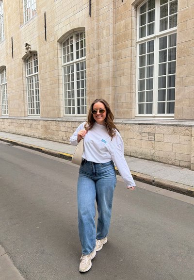 Mujer joven con gafas de sol, camisa blanca de manga larga, vaqueros azules y zapatillas caminando por una acera junto a un edificio de ladrillo beige con ventanas.