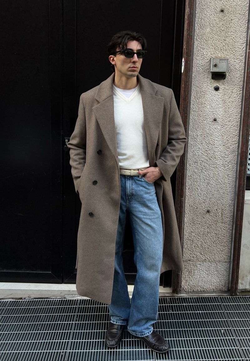 Homme portant un pardessus beige, un pull blanc, un jean bleu, des chaussures noires et des lunettes de soleil, debout avec une main dans la poche près d'un mur texturé.