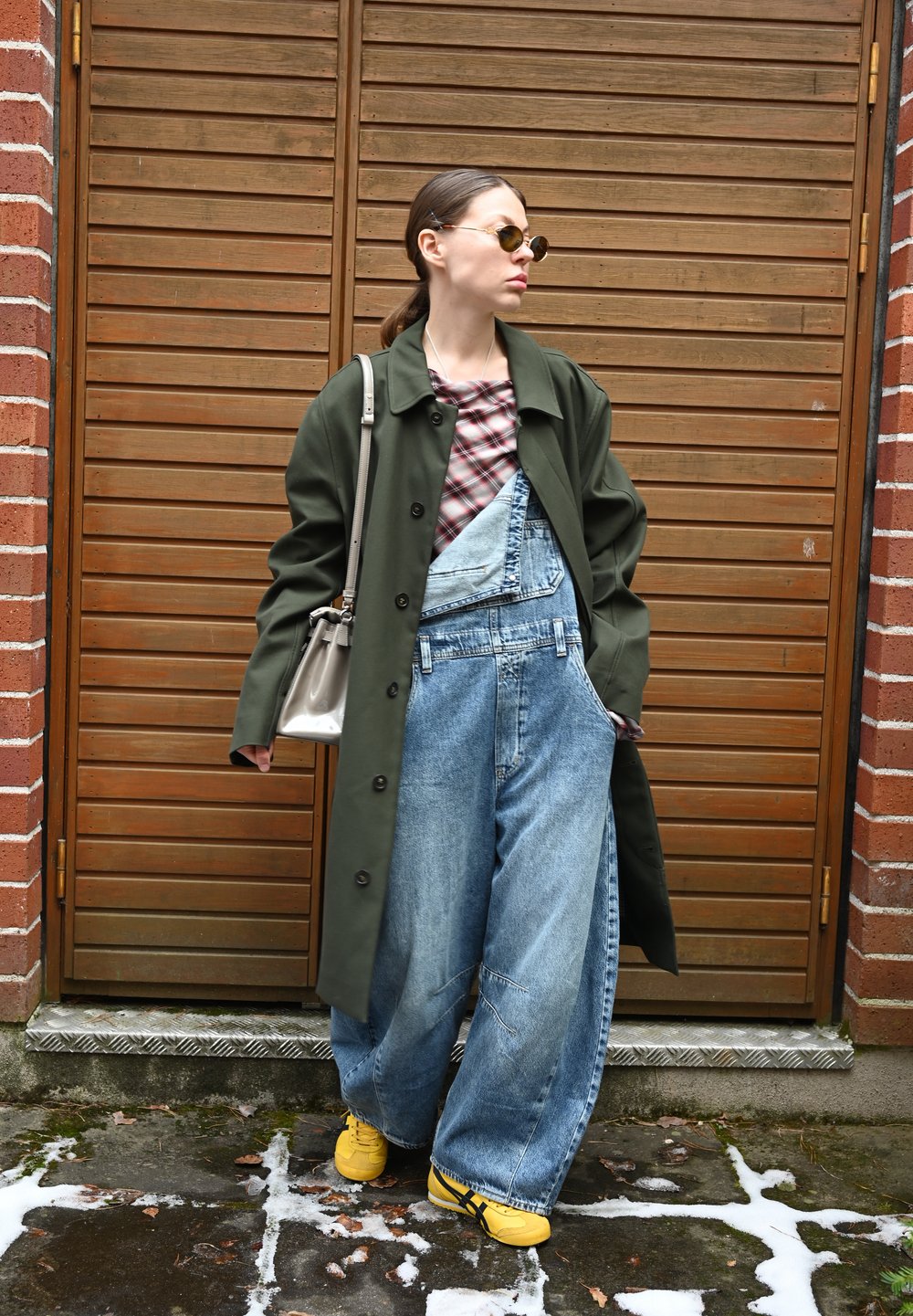 Vrouw in oversized denim overall, groene jas, gele sneakers en zonnebril die tegen een houten deur met bakstenen muren staat.