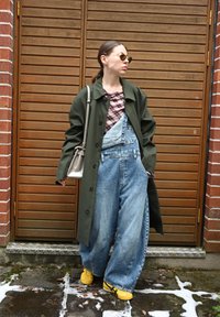 Vrouw in oversized denim overall, groene jas, gele sneakers en zonnebril die tegen een houten deur met bakstenen muren staat.