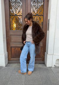 Abrigo de piel sintética marrón sobre una camiseta de manga larga a rayas, combinado con vaqueros acampanados azul claro y botas de ante marrón claro. Accesorio de bolso negro. Fondo de puerta de madera.