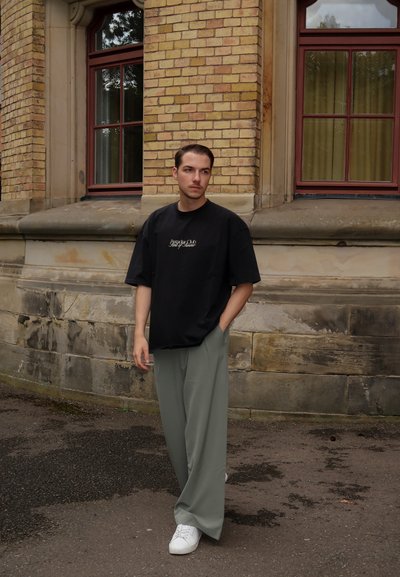 Camiseta negra de gran tamaño con logotipo, pantalones anchos verde claro y zapatillas blancas. El fondo presenta una pared de ladrillo y grandes ventanas.