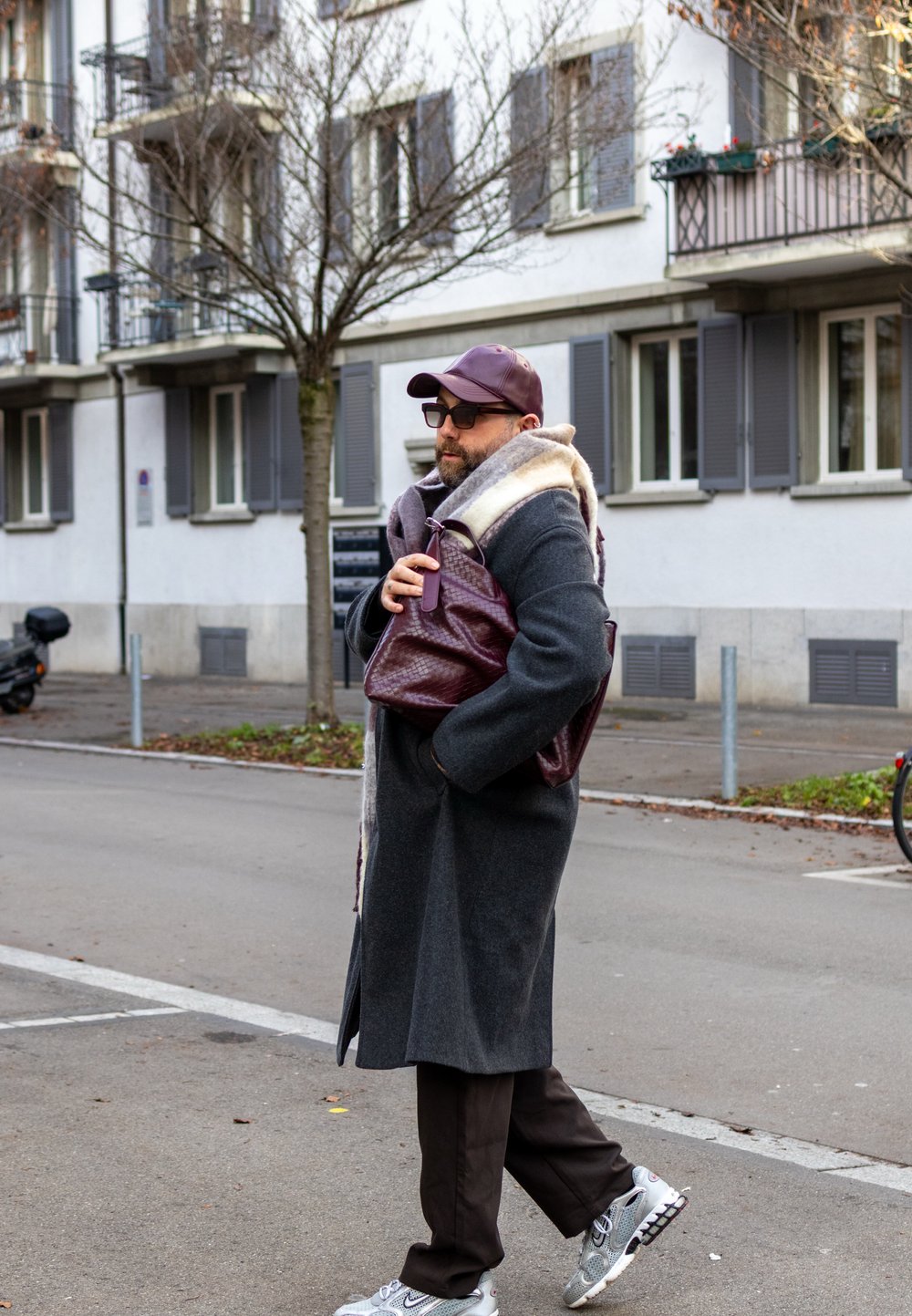 Harmaa päällystakki, jossa vaalea turkisvuori, ruskeat housut ja harmaat tennarit. Kantaa violettia tekstuuripintaista laukkua. Kaupungin ympäristö, taustalla puu.