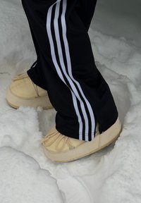 Pieds portant des bottes d'hiver beiges et un pantalon noir avec des rayures blanches, debout dans une neige profonde.