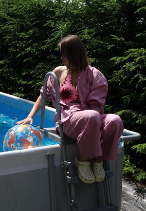 Mujer en pijama rosa y zapatillas crema se sienta en la escalera de una piscina elevada, tocando una pelota inflable con un patrón de peces coloridos en el agua.