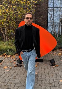 Blazer nero sopra un dolcevita nero, jeans azzurri e occhiali da sole; in piedi accanto a una grande forma astratta arancione brillante sul marciapiede.
