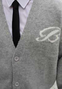 Halli kootud kardigan v-kaelusega, millel on valge täht "B" ja lillakad nupud, kantud helelilla särgi ja musta lipsu peal.