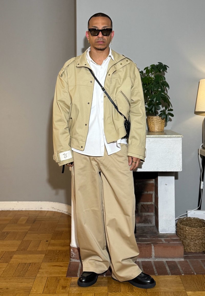 Homme portant une veste beige et un pantalon large, chemise blanche, chaussures noires, lunettes de soleil et sac en bandoulière, debout à l'intérieur près d'une petite cheminée et d'une plante.