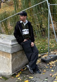 Mężczyzna w czarnej bluzie z kapturem na zamek, zielonym polo z białymi paskami, ciemnych dżinsach i szarych trampkach, oparty o kamienną strukturę.