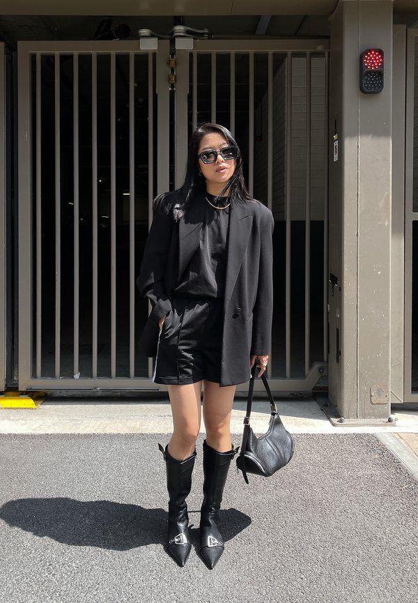Blazer preto sobre uma blusa preta, combinado com calções pretos. Botas de salto agulha pretas até ao joelho e uma pequena carteira preta. Óculos de sol estão a ser usados.