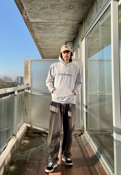 Sudadera con capucha gris claro con el logo "carhartt", pantalones gris oscuro con franjas laterales y zapatillas negras. Fondo de un balcón con una separación de vidrio texturizado.
