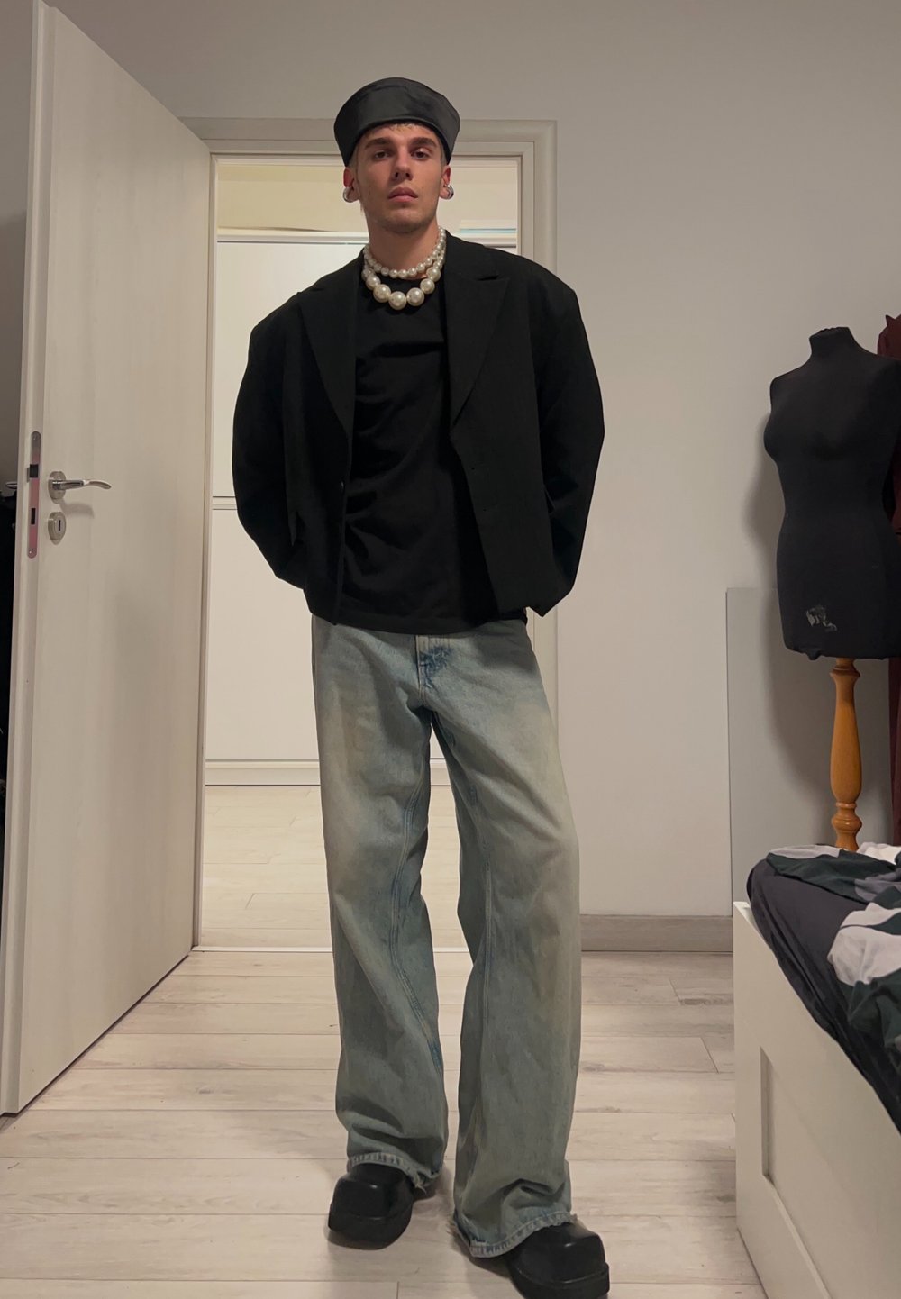 Zwarte blazer over een zwart t-shirt, lichtblauwe wijdlopende jeans, zwarte schoenen en een zwarte pet. Parelsnoer en zachte texturen.