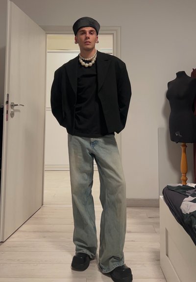 Blazer negro sobre una camiseta negra, jeans de pierna ancha azul claro, zapatos negros y una gorra negra. Collar de perlas y texturas suaves.