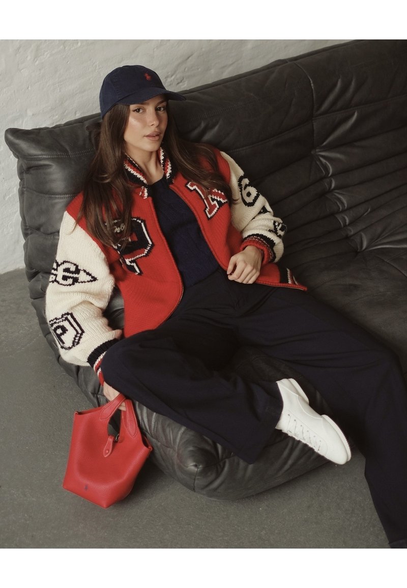 Young woman reclining on a dark leather sofa wearing a red and cream varsity jacket, navy trousers, white trainers, navy cap, and holding a red handbag.