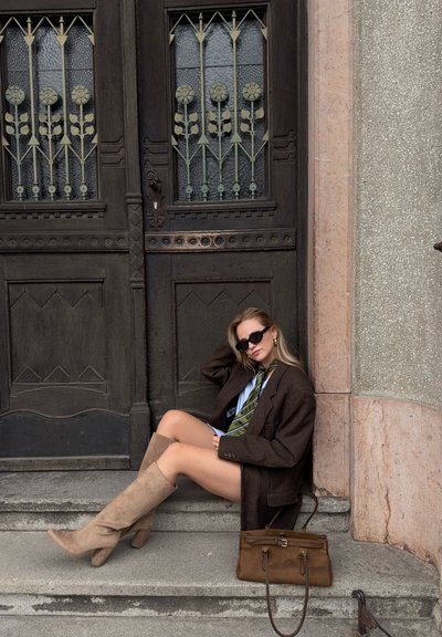 Blazer oversized de cuadros marrones, camisa azul con corbata verde, botas de rodilla beige, bolso marrón; sentado contra una puerta de madera decorativa.