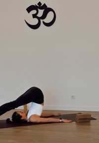 Personne en haut noir et leggings démontrant une posture de yoga sur un tapis, avec des blocs en liège à proximité et un symbole "Om" sur le mur.