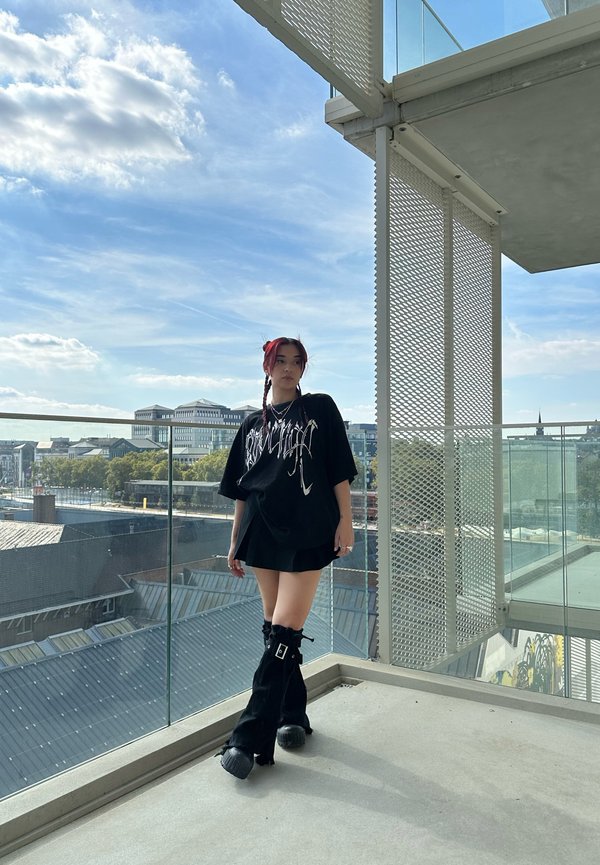 Overmaat zwarte graphic t-shirt met witte tekst, laag over een zwarte mini-jurk. Zwarte kniehoge laarzen met een gespdetail. Stedelijke omgeving, blauwe lucht.