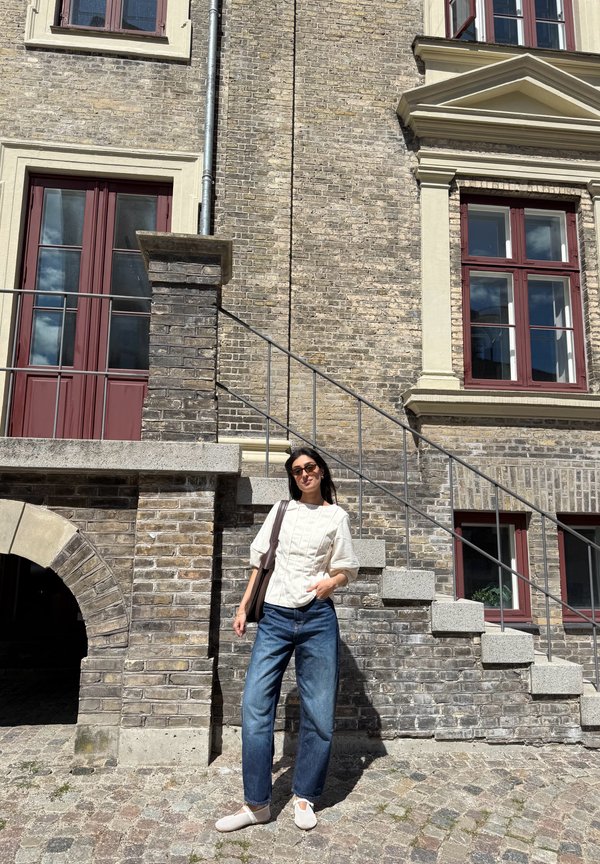 Femme portant des lunettes de soleil, un top blanc et un jean bleu, debout dans une rue pavée à côté d'un bâtiment en briques avec des fenêtres à cadre rouge et un escalier en pierre.