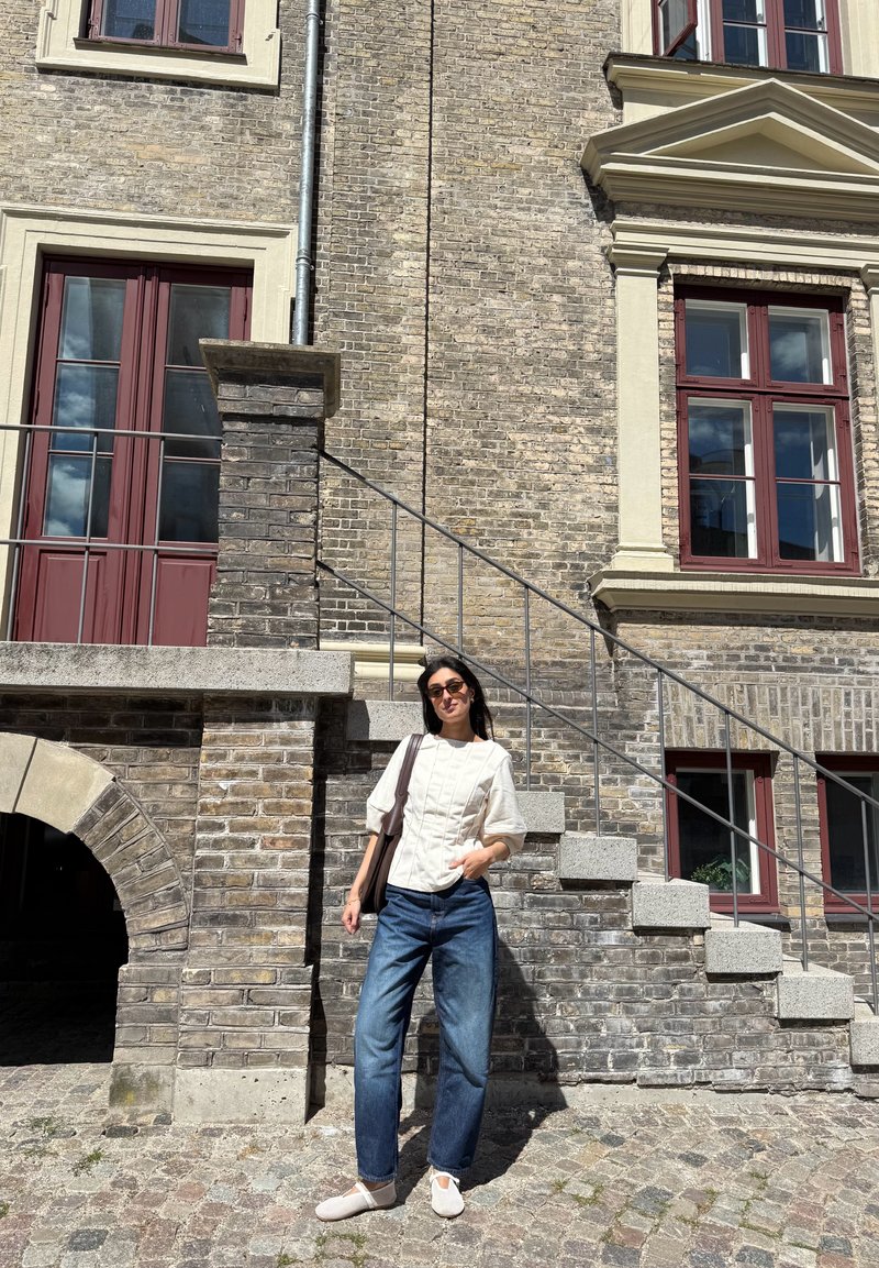 Femme portant des lunettes de soleil, un haut blanc et un jean bleu, debout dans une rue pavée à côté d'un bâtiment en brique avec des fenêtres à cadres rouges et un escalier en pierre.