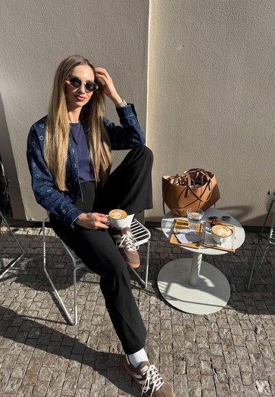 Mujer sentada en una silla blanca, vistiendo una chaqueta de mezclilla con patrones de corazones, pantalones negros y zapatillas. Sosteniendo una taza de café sobre una mesa.
