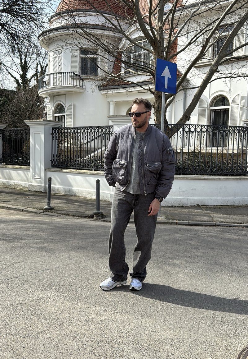 Homme portant des lunettes de soleil et une veste grise debout dans une rue devant un bâtiment blanc avec une clôture noire et un panneau de sens unique.