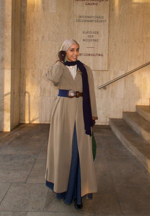 Mujer sonriente con gabardina beige, bufanda azul, guantes y hiyab posa cerca de unos escalones con texto de galería de arte en la pared de piedra detrás de ella.