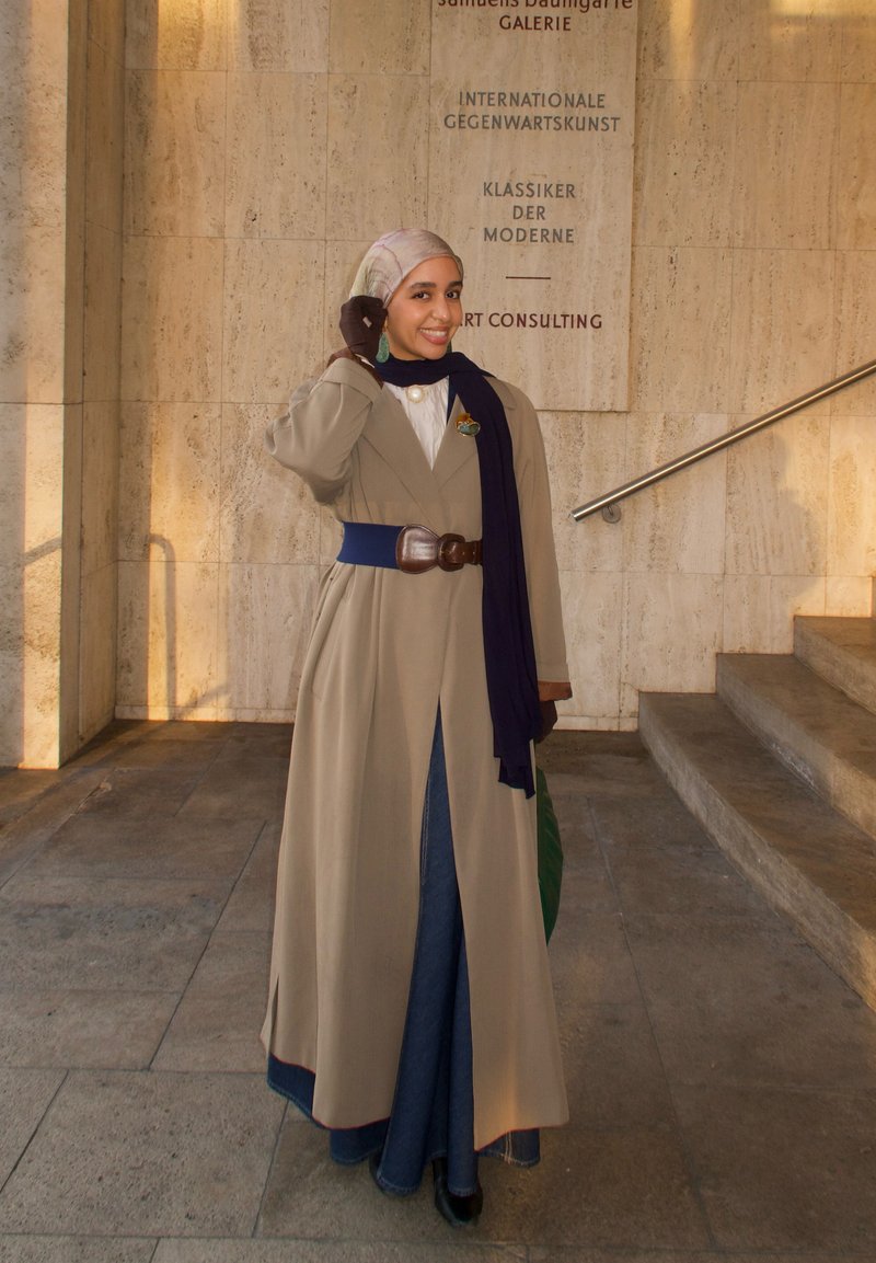 Femme souriante en trench beige, écharpe bleue, gants et hijab posant près des marches avec le texte galerie d'art sur le mur en pierre derrière elle.