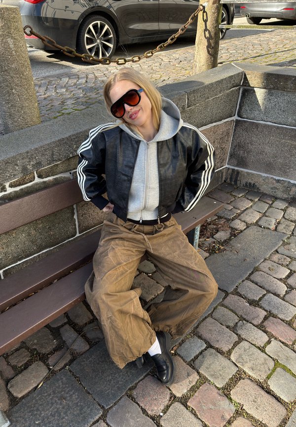 Joven mujer con gafas de sol, chaqueta negra, sudadera con capucha gris y pantalones marrones holgados sentada en un banco en una calle empedrada.