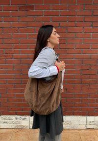 Νεαρή γυναίκα με γκρι και λευκά ρούχα που κρατά μια μεγάλη καφέ σακούλα ώμ ου από σουέτ, στέκεται πλευρικά δίπλα σε έναν τοίχο από κόκκινα τούβλα.