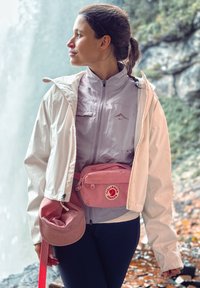 Rosa fanny pack med logo, lett grå jakke, blå topp og rød aksentstropp. Akkompagnert av en rosa bøttehatt med teksturert stoff.